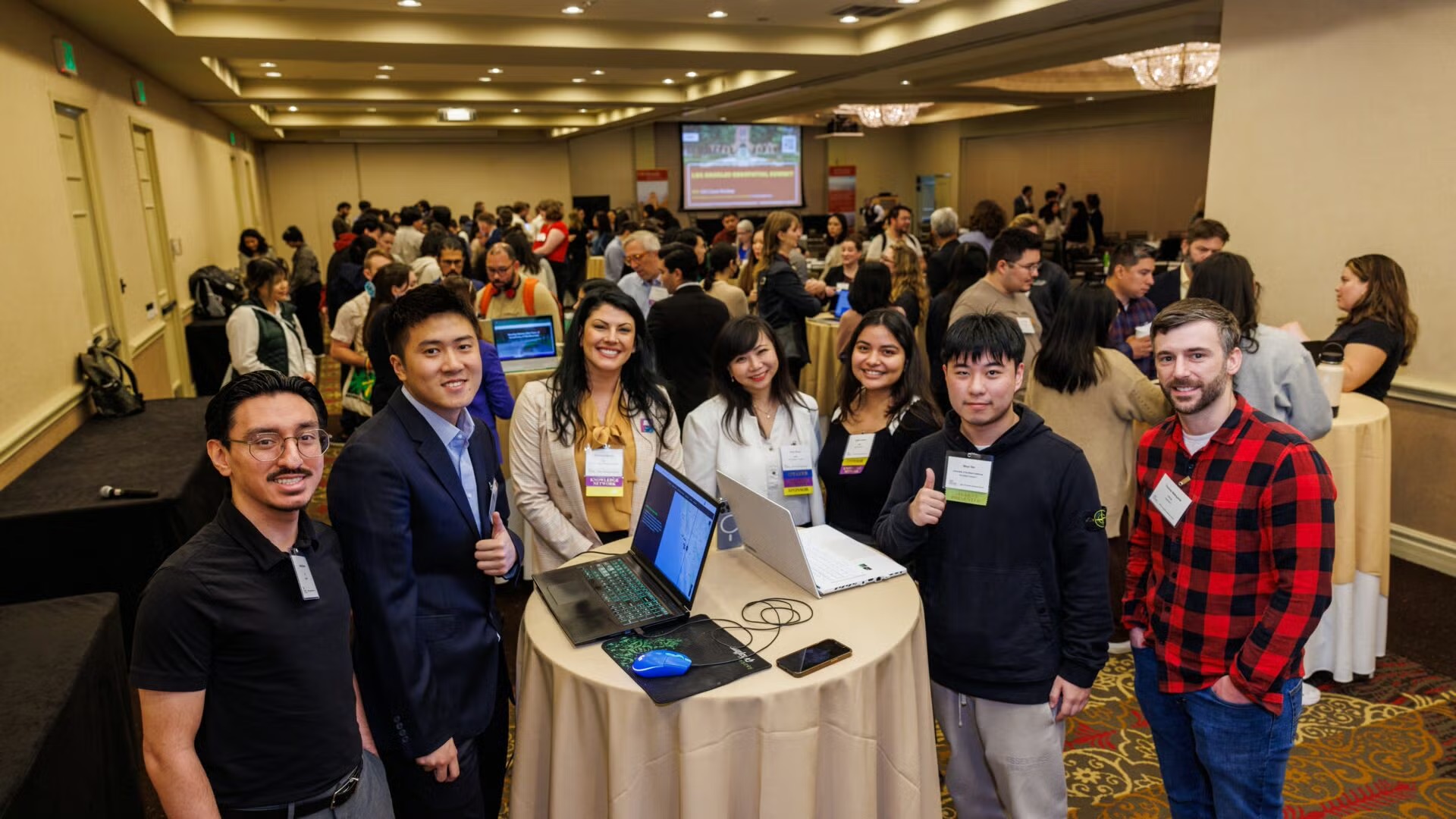two males adn two females standing around a table with laptops open smiling